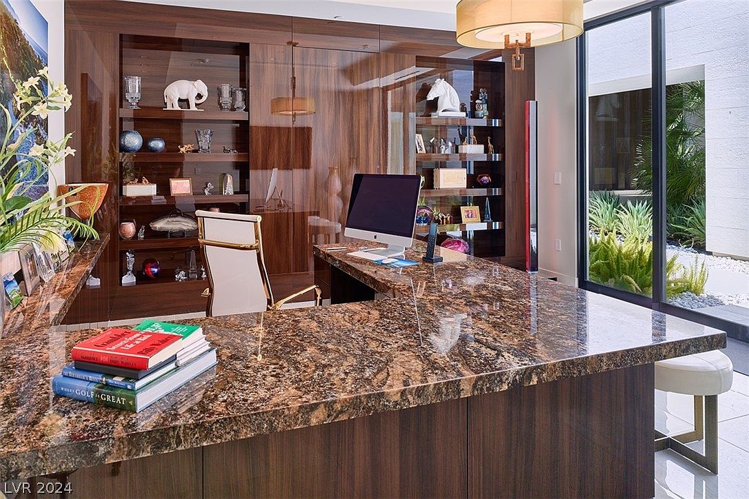 This is an interior shot of a luxurious home office. The room features a large, brown granite desk, custom wood built-in shelving with decorative items, and a modern chair. A large window provides natural light and a view of the exterior landscaping, creating a sophisticated and productive workspace.