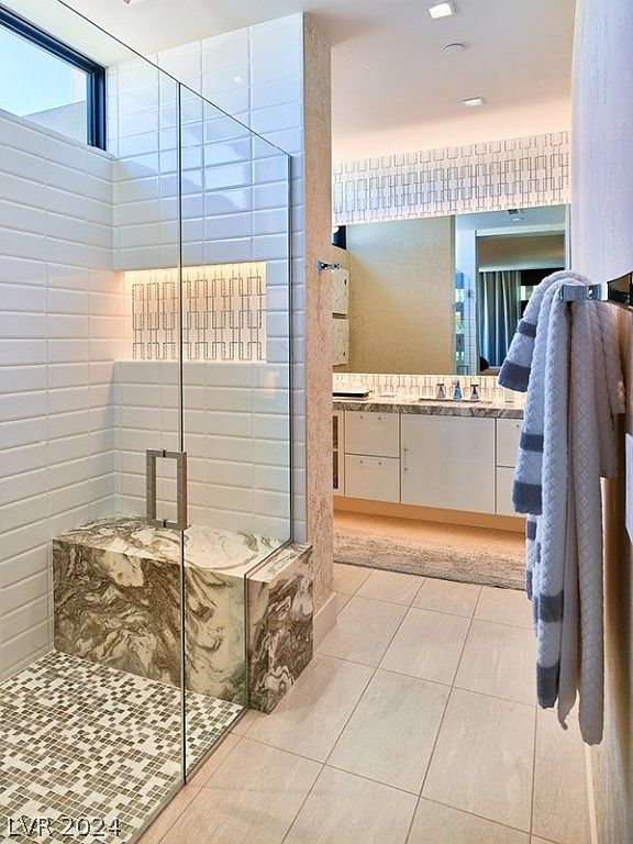 This is a primary bathroom featuring a glass-enclosed shower with a marble bench and mosaic tile floor. The bathroom has a modern design with white subway tiles, a large mirror, and a vanity with white cabinets and marble countertops. A plush rug and neatly hung towels add a touch of luxury.