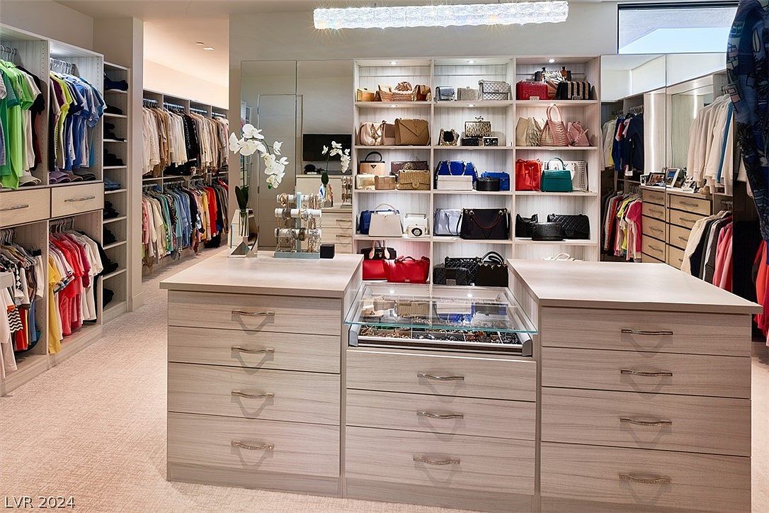 This is a well-organized walk-in closet featuring custom shelving and drawers. The closet is filled with clothing, handbags, and accessories, all neatly arranged. A central island with a glass-topped jewelry display adds a luxurious touch, and the overall impression is one of sophistication and ample storage.