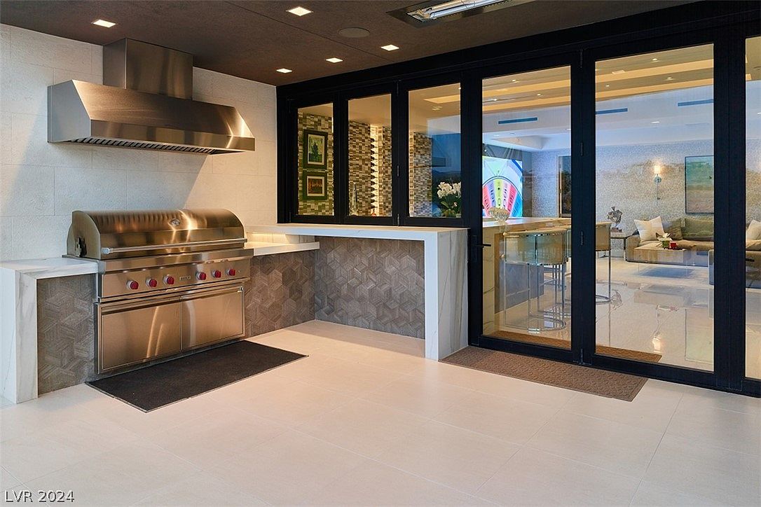 This image showcases an outdoor kitchen area, seamlessly blending indoor and outdoor living. The space features a stainless steel grill with a hood, a countertop with geometric tile accents, and a large sliding glass door that opens to the interior. The flooring is light-colored tile, and the overall design is modern and luxurious.