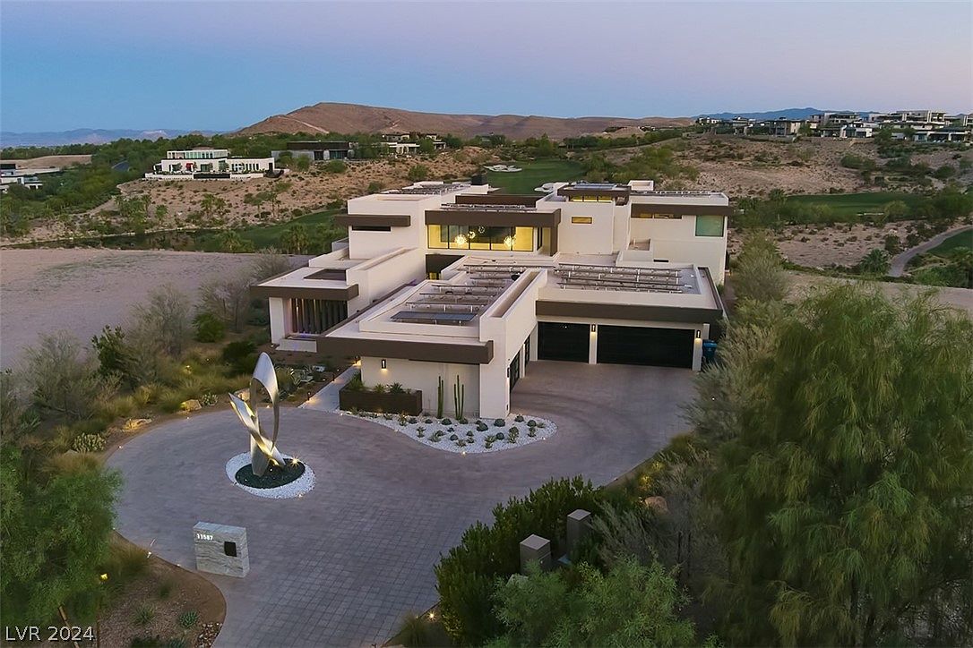This aerial view showcases a modern, multi-level home with a flat roof and solar panels. The property features a circular driveway with a sculpture, manicured landscaping, and a two-car garage. The house is situated on a hillside with views of the surrounding landscape and other luxury homes.