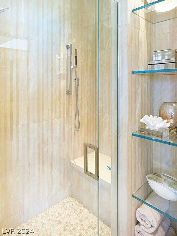 This is a well-lit primary bathroom featuring a glass-enclosed shower with a built-in bench and a handheld showerhead. The walls are covered in light-colored, vertically striped tiles, and there are glass shelves displaying decorative items and towels. The overall impression is clean, modern, and luxurious.