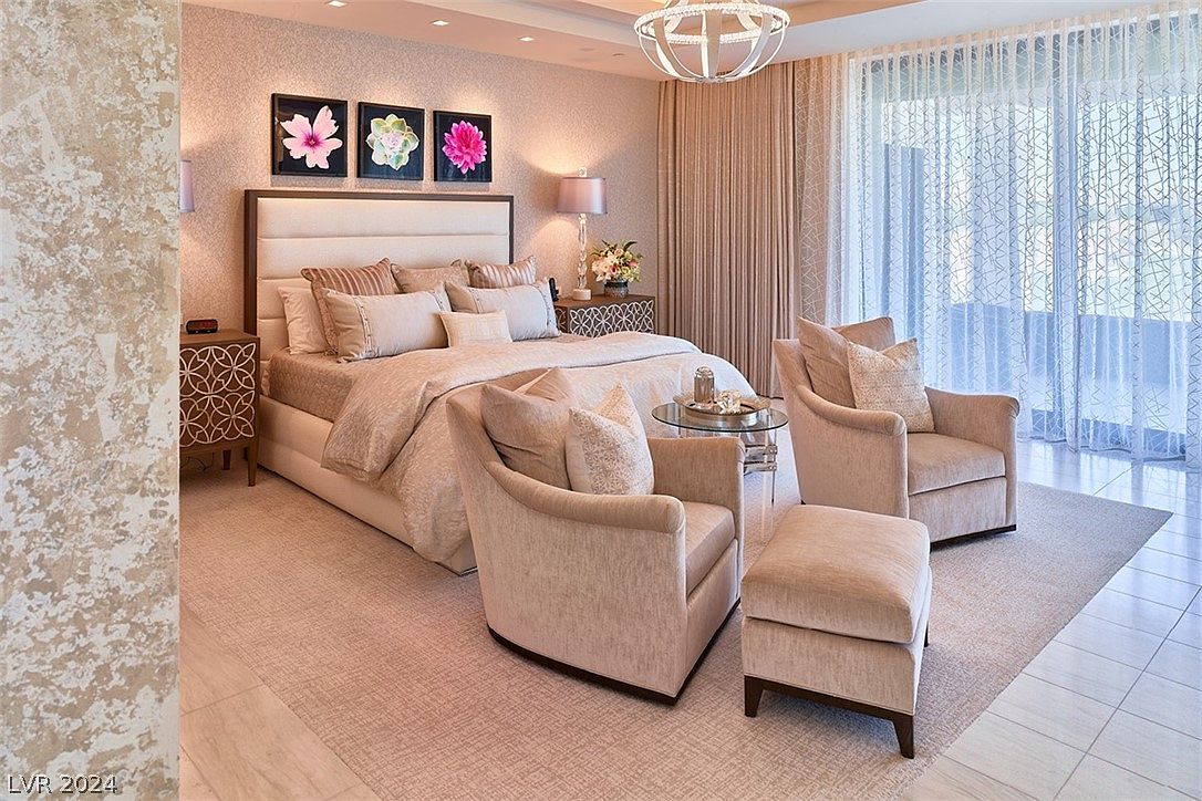 This is a luxurious primary bedroom featuring a large upholstered headboard, plush bedding, and a neutral color palette. Two comfortable armchairs and an ottoman create a cozy seating area near the window, which is covered with sheer curtains. The room is well-lit and exudes a sense of calm and sophistication.