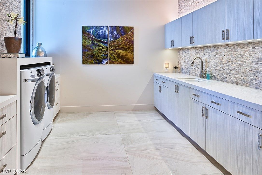 This is a well-organized laundry room featuring a front-loading washer and dryer set, light blue cabinetry with modern hardware, and a countertop with a sink. The backsplash is a mosaic tile, and the flooring is a light-colored tile with subtle veining. Artwork adorns the wall, adding a touch of personality to the space.