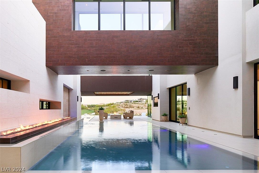 This image showcases a luxurious outdoor pool area, seamlessly integrated with the architecture of the home. The pool reflects the surrounding structure, including a striking brick facade and large windows. A linear fire feature adds warmth and ambiance, while the open design provides views of the landscape beyond, creating a serene and sophisticated atmosphere.