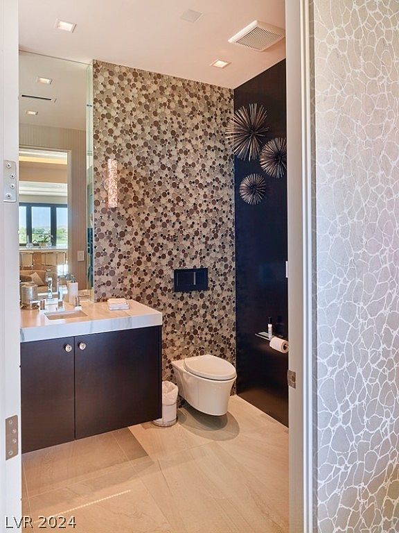 This is a modern guest bathroom featuring a floating vanity with dark wood cabinets and a white countertop. The wall behind the toilet is decorated with a unique circular mosaic tile pattern, while the adjacent wall is a dark color with decorative metal accents. The flooring is a light-colored tile, and the overall design is sleek and contemporary.