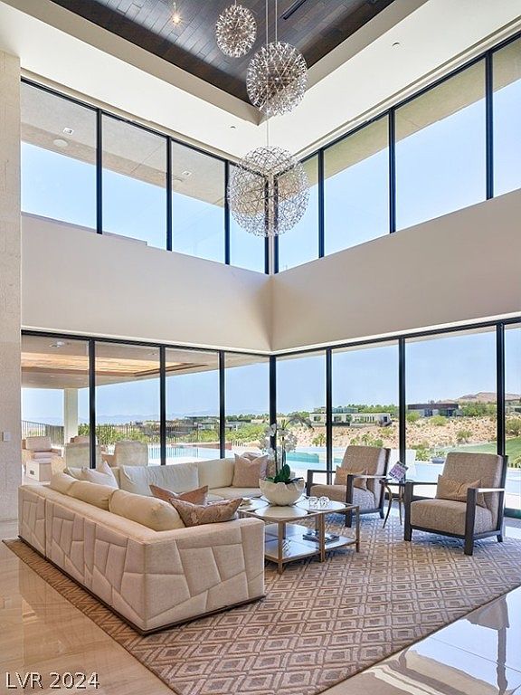 This is a luxurious living room featuring floor-to-ceiling windows that offer expansive views of the landscape. The room is furnished with a modern, geometrically patterned sofa, two armchairs, and a stylish coffee table, all set on a patterned rug. Three spherical chandeliers hang from the high ceiling, adding a touch of elegance and drama to the space.