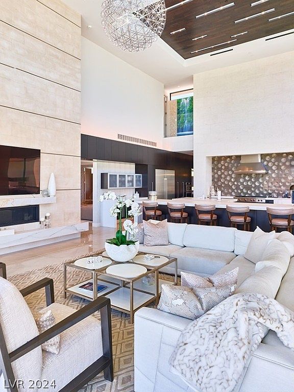 This is a luxurious living room featuring a high ceiling, a modern fireplace, and a large sectional sofa. The room is decorated in neutral tones with pops of gold, creating a sophisticated and inviting atmosphere. The open floor plan seamlessly connects the living room to the kitchen area, enhancing the sense of spaciousness.