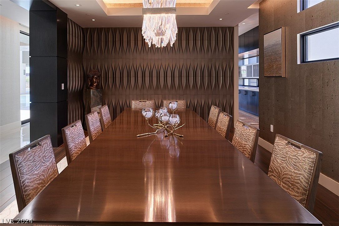 This is an interior shot of a dining room featuring a long, polished wooden table with seating for many. A modern chandelier hangs above the table, and a decorative centerpiece adds to the room's elegance. The walls are adorned with textured panels, and a glimpse into an adjacent room suggests an open floor plan, creating a sophisticated and inviting atmosphere.