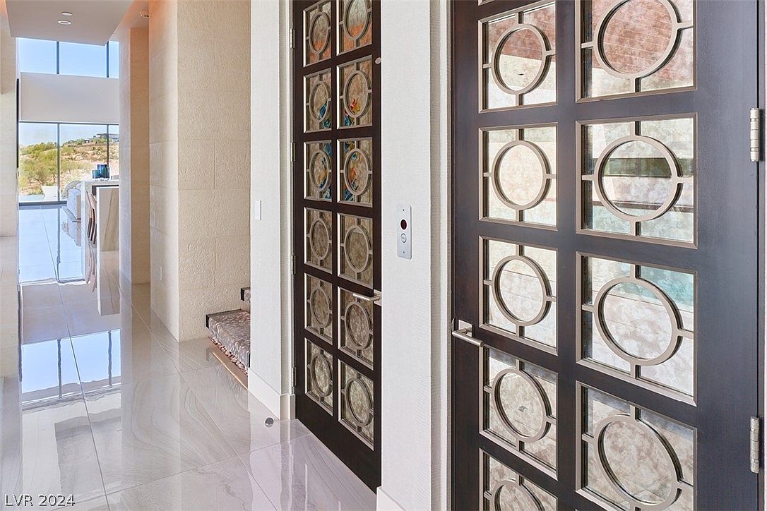 This interior shot showcases a modern hallway with high-gloss tile flooring that reflects natural light from a large window at the end of the hall. Two decorative doors with intricate circular designs flank the hallway, adding a touch of elegance and visual interest. The neutral color palette and clean lines contribute to a sophisticated and spacious feel, ideal for a luxury real estate listing.