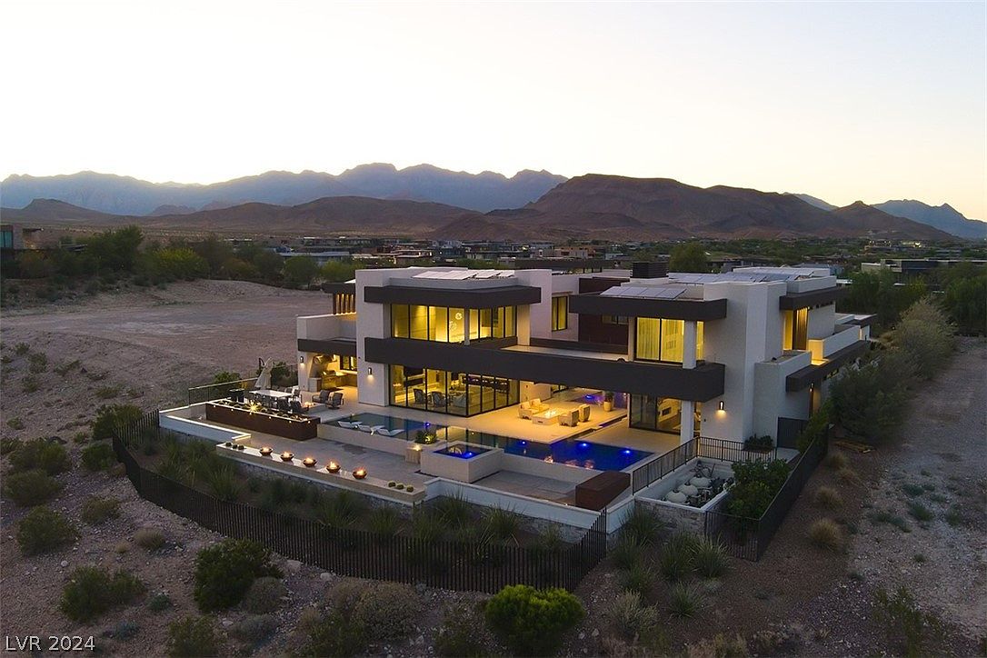 This aerial view showcases a modern, multi-level home with a flat roof and clean architectural lines. The property features a pool, spa, and outdoor entertaining areas, all set against a backdrop of mountains. The landscape is desert-like, providing a sense of privacy and exclusivity.