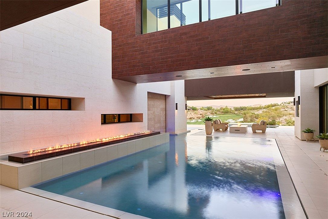 This image showcases a luxurious outdoor pool area with a modern architectural design. The pool is surrounded by sleek, light-colored tiling and features a linear fire element along one side. The backdrop includes a well-manicured golf course and desert landscape, visible through an open-air structure with comfortable outdoor seating, creating a seamless indoor-outdoor living experience.
