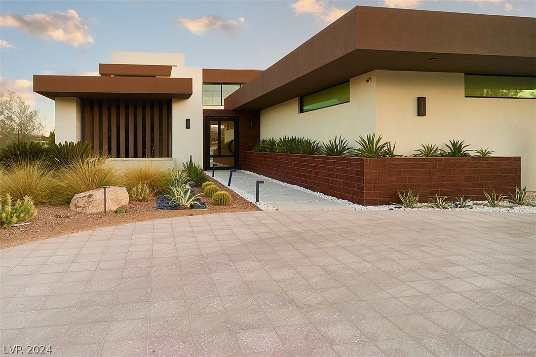 This is a front exterior view of a modern, single-story home. The house features a flat roof with brown accents, a light-colored stucco exterior, and a contemporary front door. The landscaping includes desert plants, decorative rocks, and a paved driveway, creating a clean and stylish curb appeal.