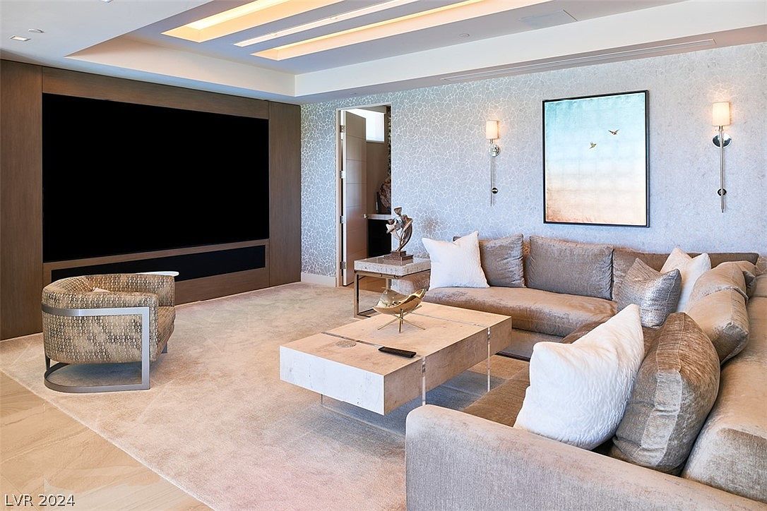 This is an interior shot of a luxurious living room featuring a large sectional sofa with decorative pillows, a modern coffee table, and a built-in entertainment center with a large screen. The room is decorated in neutral tones with patterned wallpaper and stylish lighting fixtures, creating a sophisticated and comfortable atmosphere. The perspective is from a medium distance, capturing the entire living space.
