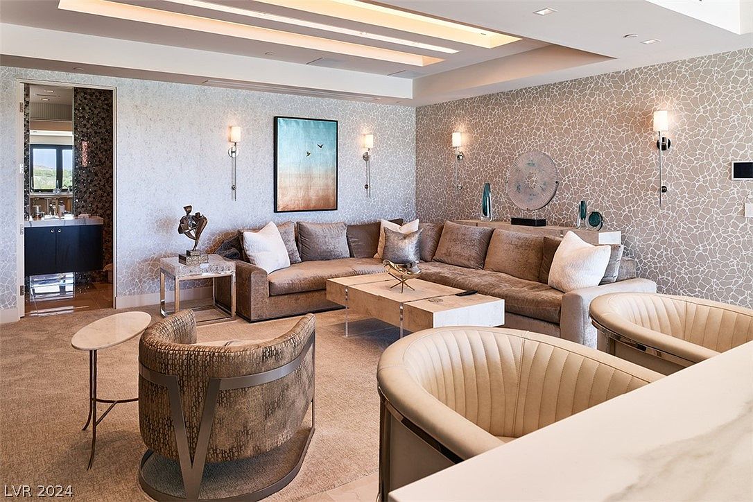 This is an interior shot of a luxurious living room featuring a large, comfortable sectional sofa, stylish armchairs, and a modern coffee table. The walls are adorned with textured wallpaper and elegant sconces, complemented by a striking piece of artwork. The room exudes a sophisticated and inviting atmosphere, perfect for relaxation and entertaining.
