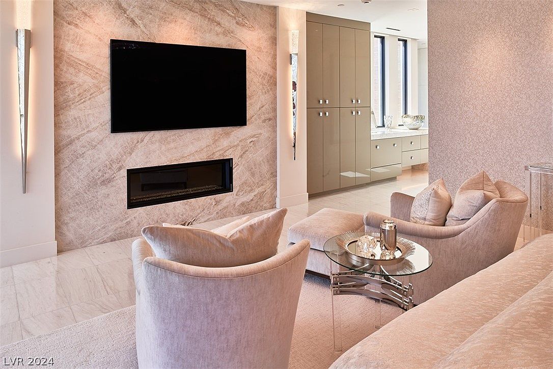 This interior shot showcases a luxurious living room with a modern fireplace and a large flat-screen TV mounted above it. Two plush armchairs and a glass-topped coffee table create an inviting seating area. The room features elegant wall finishes and flooring, contributing to a sophisticated and comfortable atmosphere.