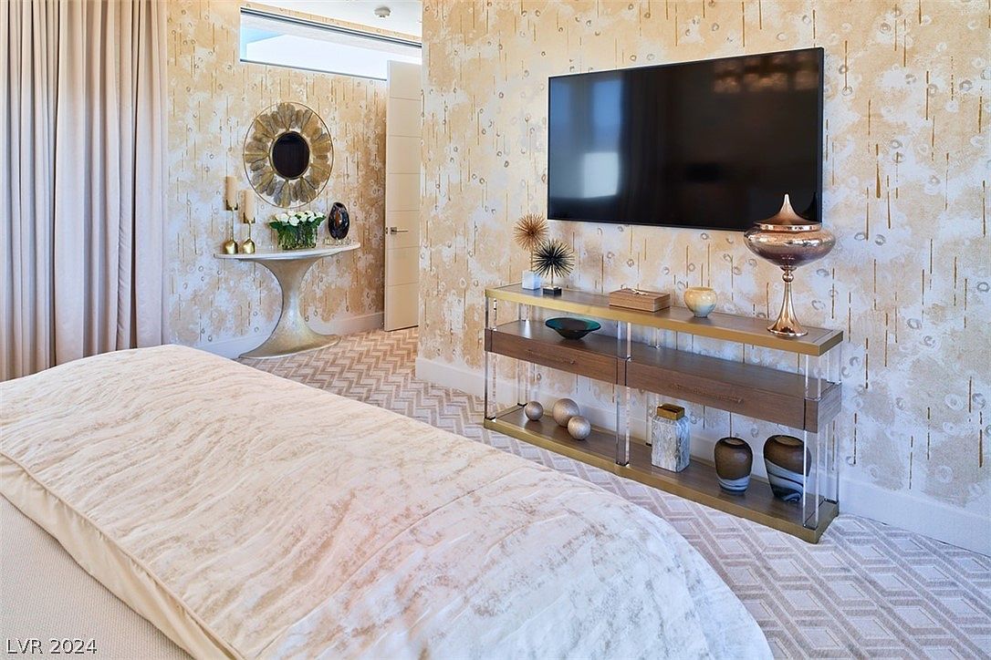 This is a primary bedroom featuring a large bed with a textured white comforter. The room has patterned wallpaper and a console table with a large flat-screen television mounted above it. A round mirror and decorative table are visible in the background, adding to the room's luxurious feel.