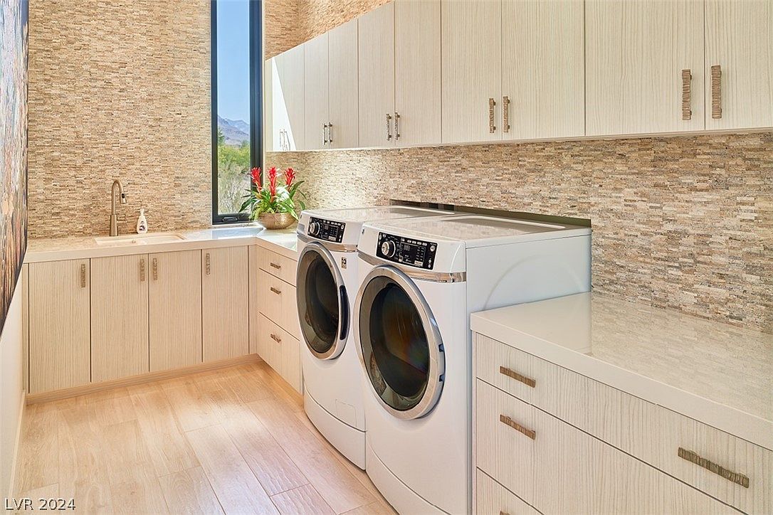 This is a well-appointed laundry room featuring a modern washer and dryer set, light wood cabinetry with sleek hardware, and a countertop that extends over the machines and sink area. The backsplash and adjacent wall are adorned with a textured, neutral-toned tile, adding visual interest. A window provides natural light and a view to the outside, enhancing the room's appeal.
