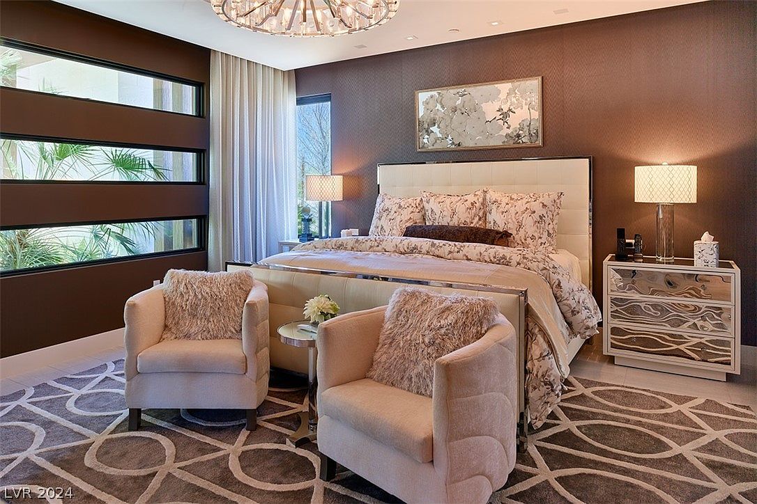 This is a luxurious primary bedroom featuring a large bed with a tufted headboard and floral bedding. Two plush armchairs flank a small table, and a mirrored nightstand sits beside the bed. The room is decorated in warm brown tones with a patterned rug, creating a cozy and elegant atmosphere.