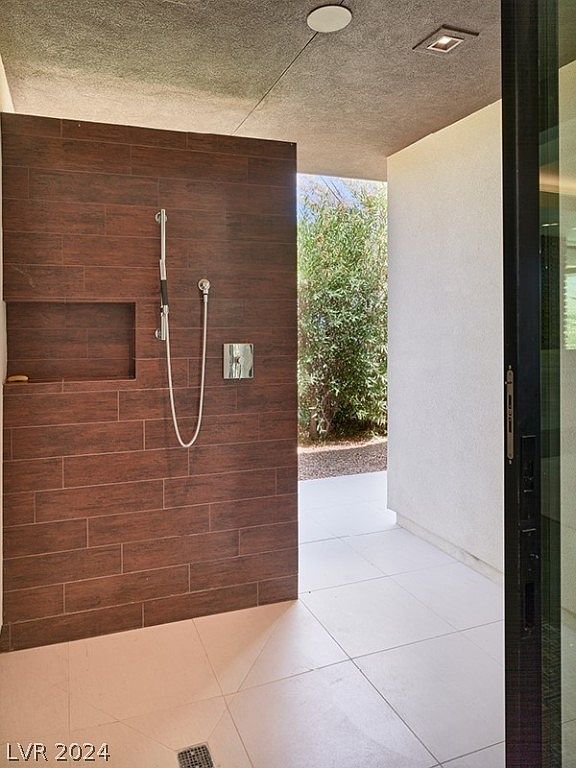 This image showcases a modern primary bathroom with a unique open shower design. The shower features wood-look tile walls, a built-in niche, and sleek fixtures. The flooring consists of large white tiles, and natural light filters in from the adjacent outdoor area, creating a spa-like atmosphere.