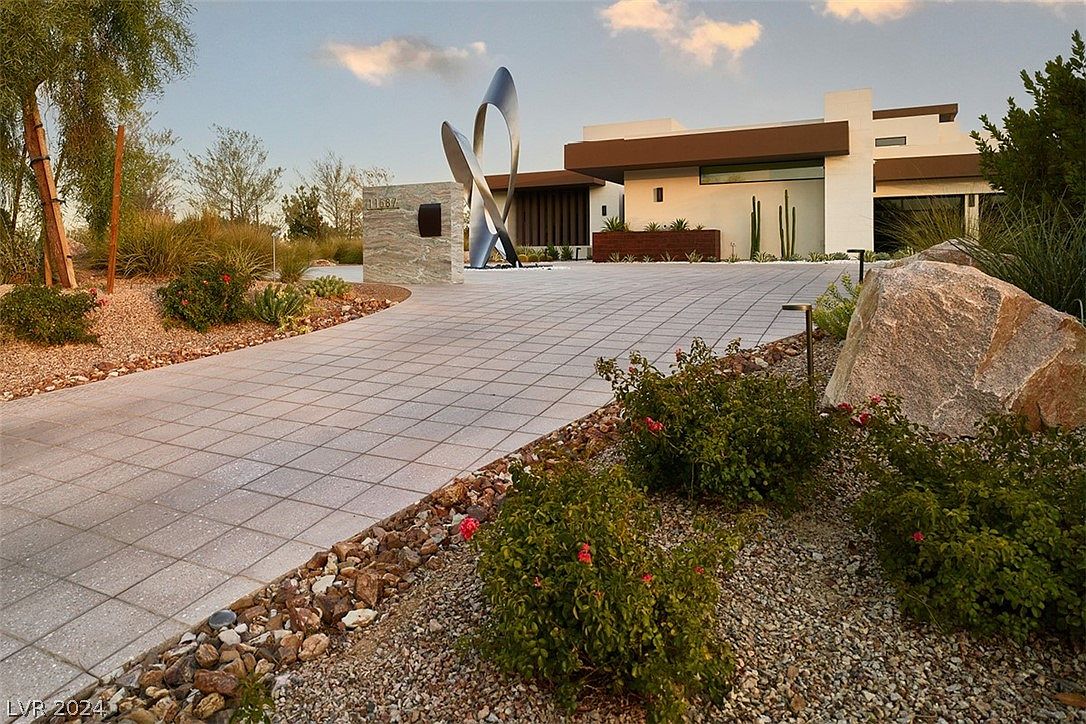 This is a front exterior view of a modern home showcasing a meticulously landscaped front yard and a contemporary architectural design. The property features a paved driveway leading to the house, complemented by decorative rocks, bushes, and a large boulder. A striking metal sculpture and a stone address marker add unique artistic elements to the entrance, enhancing the property's curb appeal.