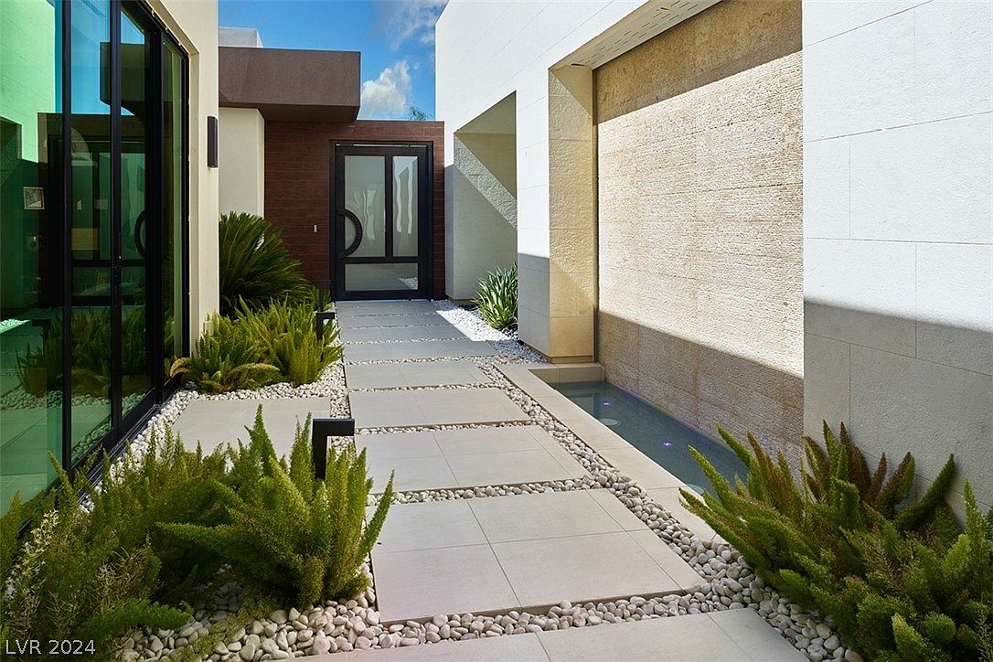 This image showcases a modern home's entryway, featuring a pathway of large square pavers separated by white pebbles, leading to a sleek, dark-framed glass door. Lush green plants line the walkway, contrasting with the minimalist architecture of the building, which includes a water feature along a textured wall. The overall impression is one of contemporary elegance and tranquility.