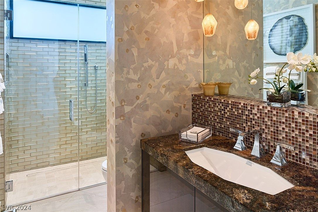 This is a luxurious primary bathroom featuring a modern vanity with a unique vessel sink and a mosaic tile backsplash. The shower is enclosed in glass and has a tiled interior. The walls have a textured finish, and pendant lights hang above the vanity, adding to the upscale ambiance.
