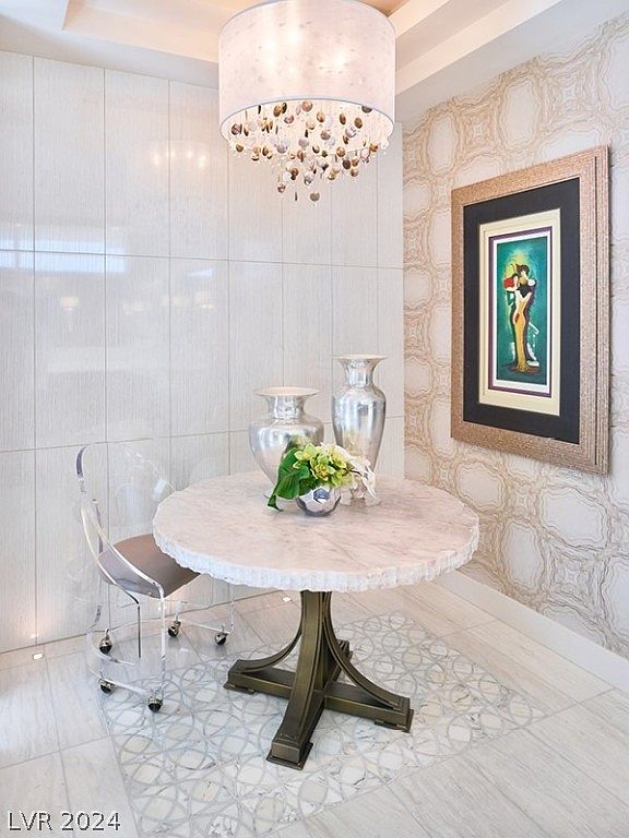 This interior shot showcases a stylish dining area featuring a round marble-topped table with a unique metal base, complemented by a modern transparent chair. Above, a decorative chandelier adds a touch of elegance, while a framed artwork adorns the patterned wall, creating a sophisticated and inviting atmosphere. The flooring features a decorative tile inlay, adding to the room's overall charm.