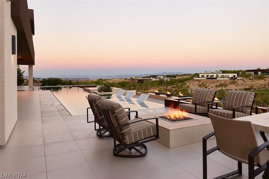 This exterior shot showcases a luxurious patio area with a modern fire pit surrounded by comfortable seating. A sleek infinity pool extends towards a scenic landscape, creating a serene and upscale ambiance. The patio features clean lines and neutral tones, enhancing the sophisticated outdoor living space.