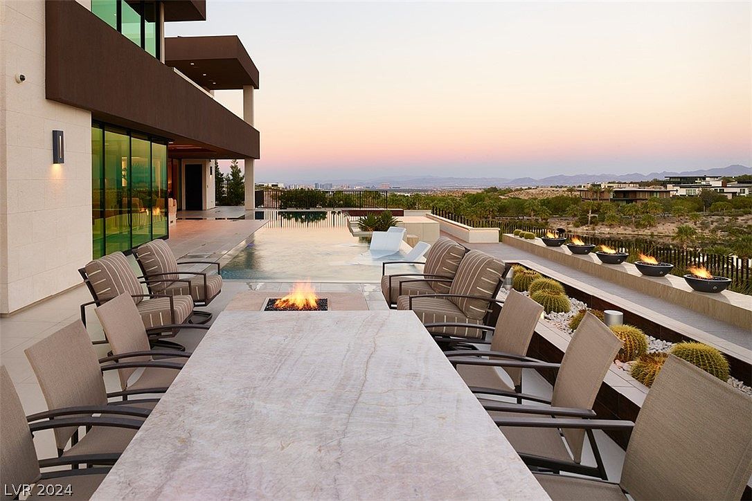 This is a luxurious outdoor patio featuring a large dining table with chairs, a fire pit, and comfortable lounge seating. The patio overlooks a stunning view of the landscape and a modern pool. The design incorporates fire features and desert landscaping, creating a sophisticated and inviting outdoor living space.