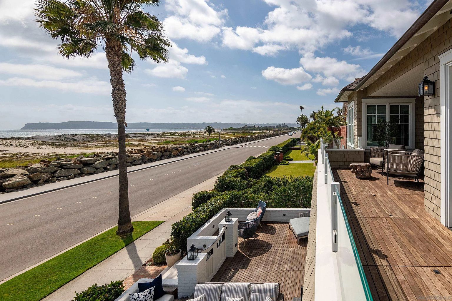This elevated perspective captures a luxurious multi-level outdoor living space featuring a wooden deck with comfortable lounge seating and a glass railing. The scene overlooks a quiet coastal road, a rocky shoreline, and the expansive ocean beyond, framed by a tall palm tree. The architecture showcases a refined coastal aesthetic with shingle siding and clean, modern lines, creating an inviting atmosphere for relaxation.