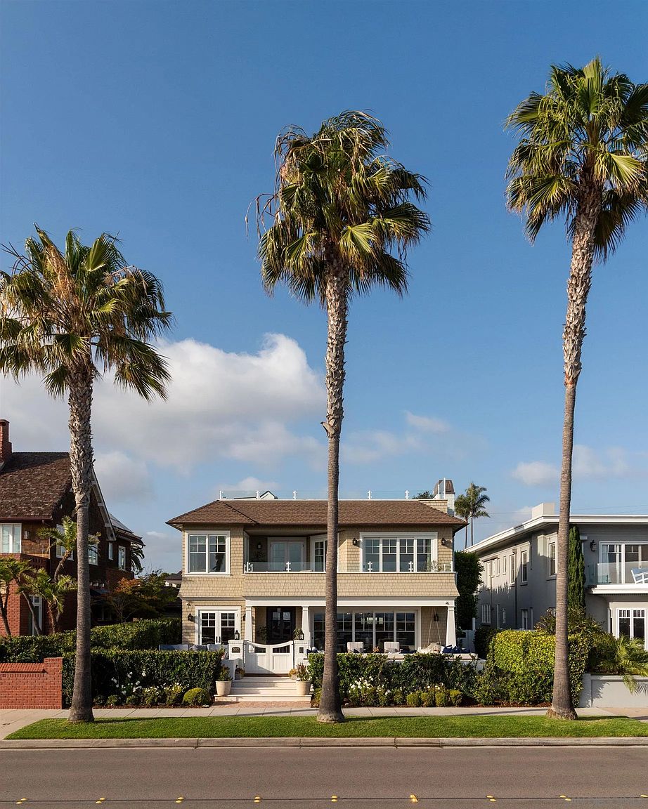 This front-facing view captures a charming two-story coastal-style home framed by three tall, mature palm trees. The residence features light-toned shingle siding, a welcoming gated entryway with a small staircase, and a second-story balcony with glass railings. A lush, manicured hedge provides privacy for the front patio area, creating a serene and inviting curb appeal under a clear blue sky.