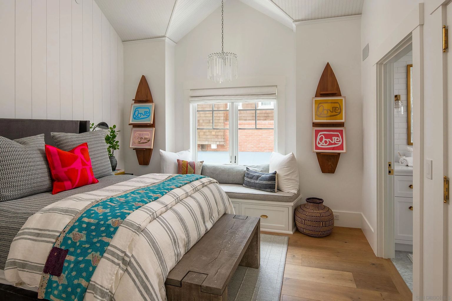 This bright and airy bedroom features a vaulted ceiling with white paneling and a central crystal chandelier. A cozy window seat with built-in storage sits beneath a large window, flanked by decorative surfboard-shaped wall art. The room is furnished with a bed dressed in patterned textiles and a rustic wooden bench at the foot, creating a relaxed, coastal-inspired atmosphere.