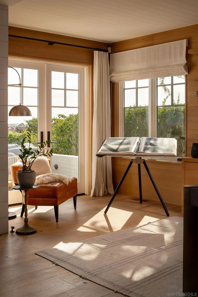 This sun-drenched study features warm wood-paneled walls and light-toned flooring, creating a serene and sophisticated atmosphere. A leather armchair with a soft throw blanket sits beside a small side table and a potted plant, while a large open book rests on a tripod stand near the window. The room is bathed in natural light from the glass doors and windows, which offer a view of the outdoor greenery.