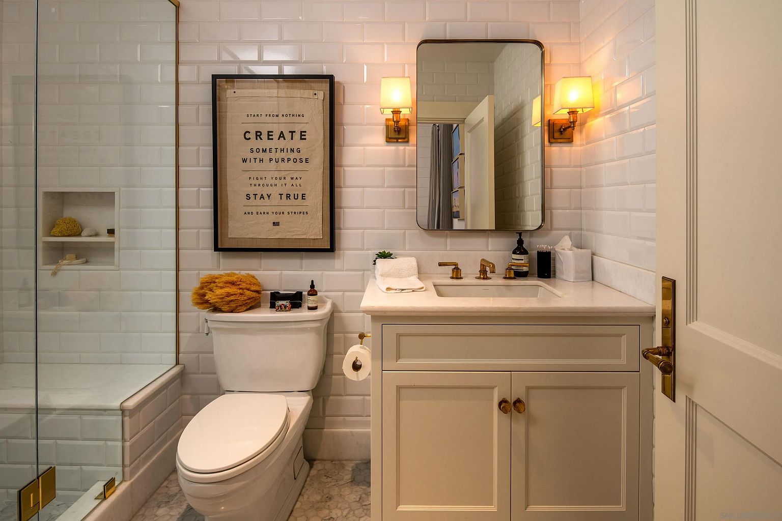 This bright and compact bathroom features white subway tile walls, a vanity with a stone countertop, and a framed mirror flanked by two wall-mounted sconces. A glass-enclosed shower area is visible to the left, while a framed inspirational print hangs above the toilet, adding a personalized touch to the space. The room is finished with patterned floor tiles and brass-toned hardware, creating a clean and cohesive aesthetic.