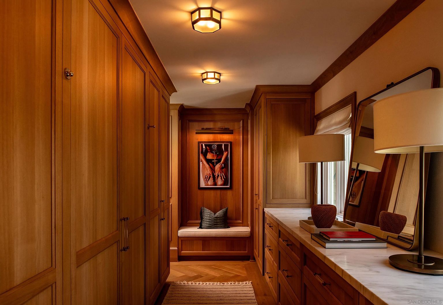 This sophisticated walk-in closet features extensive custom wood cabinetry and a built-in bench seat at the end of the hallway. A marble-topped vanity area with a large mirror and lamp provides a functional space, while warm overhead lighting enhances the rich wood tones. The perspective looks down the center of the room, highlighting the organized storage and elegant, high-end finishes.