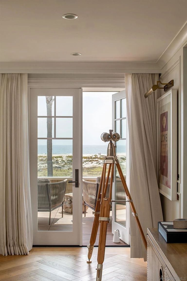 This interior view captures a sophisticated living space featuring light wood flooring arranged in a herringbone pattern. A vintage-style nautical telescope on a wooden tripod stands prominently in the foreground, framing a direct view through glass doors to a private deck and the ocean beyond. The room is accented by soft white curtains, a framed piece of art on the wall, and a warm, inviting atmosphere that seamlessly blends indoor comfort with coastal scenery.