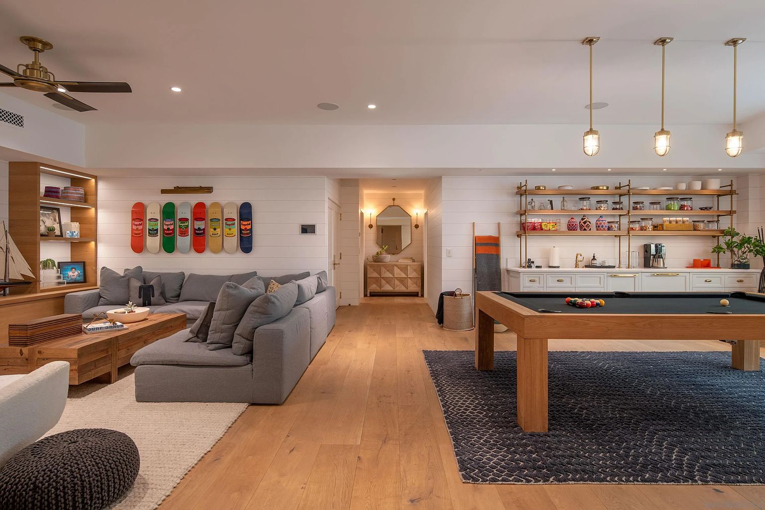 This spacious, open-concept living area features a comfortable gray sectional sofa arranged around a light wood coffee table, complemented by a pool table on a textured rug in the foreground. The room is accented by a wall display of colorful skateboard decks and a built-in shelving unit, while a wet bar with open shelving and pendant lighting sits to the right. A hallway leads to a secondary space with a mirror, creating a sense of depth and flow throughout the home.