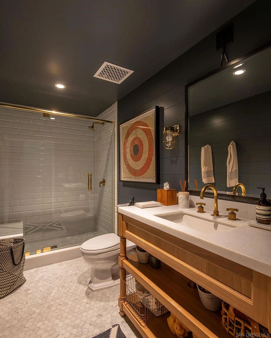 This stylish bathroom features a vanity with a light wood base, a stone countertop, and brass fixtures. The space is accented by dark-painted shiplap walls, a white subway-tiled shower enclosure with a glass door, and a classic hexagonal tile floor. A framed target print adds a unique decorative touch, creating a sophisticated and modern aesthetic.