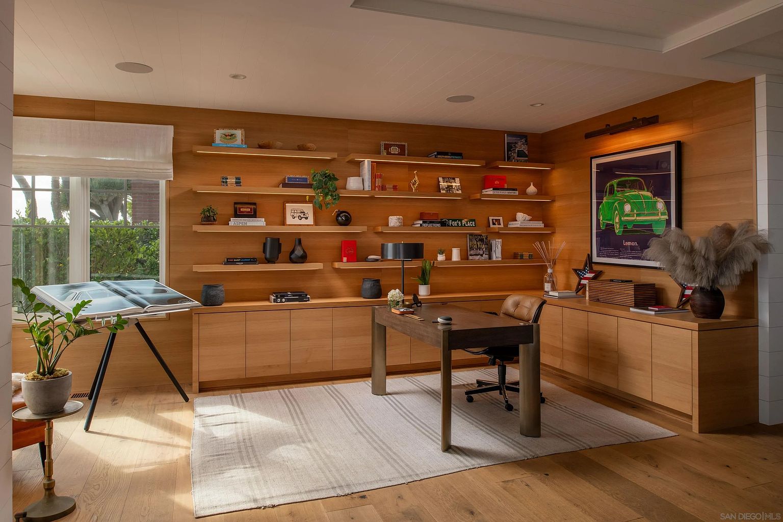 This sophisticated home office features warm, light wood paneling on the walls and custom built-in cabinetry with integrated shelving. A central desk sits atop a neutral area rug, while a large window with a roman shade allows natural light to fill the space. The room is accented by a vibrant framed print of a vintage car and decorative objects, creating a professional yet inviting atmosphere.