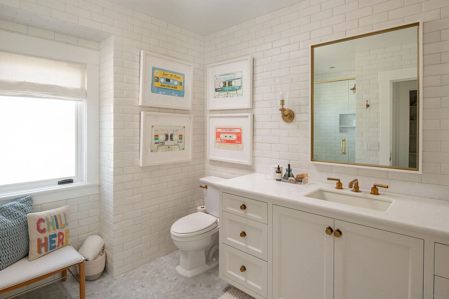 This bright and clean bathroom features white subway tile walls, a white vanity with gold hardware, and a marble-patterned floor. The space is decorated with framed cassette tape art and a bench with a decorative pillow, creating a playful yet sophisticated atmosphere. A large mirror with a gold frame hangs above the vanity, reflecting the shower area and enhancing the sense of space.