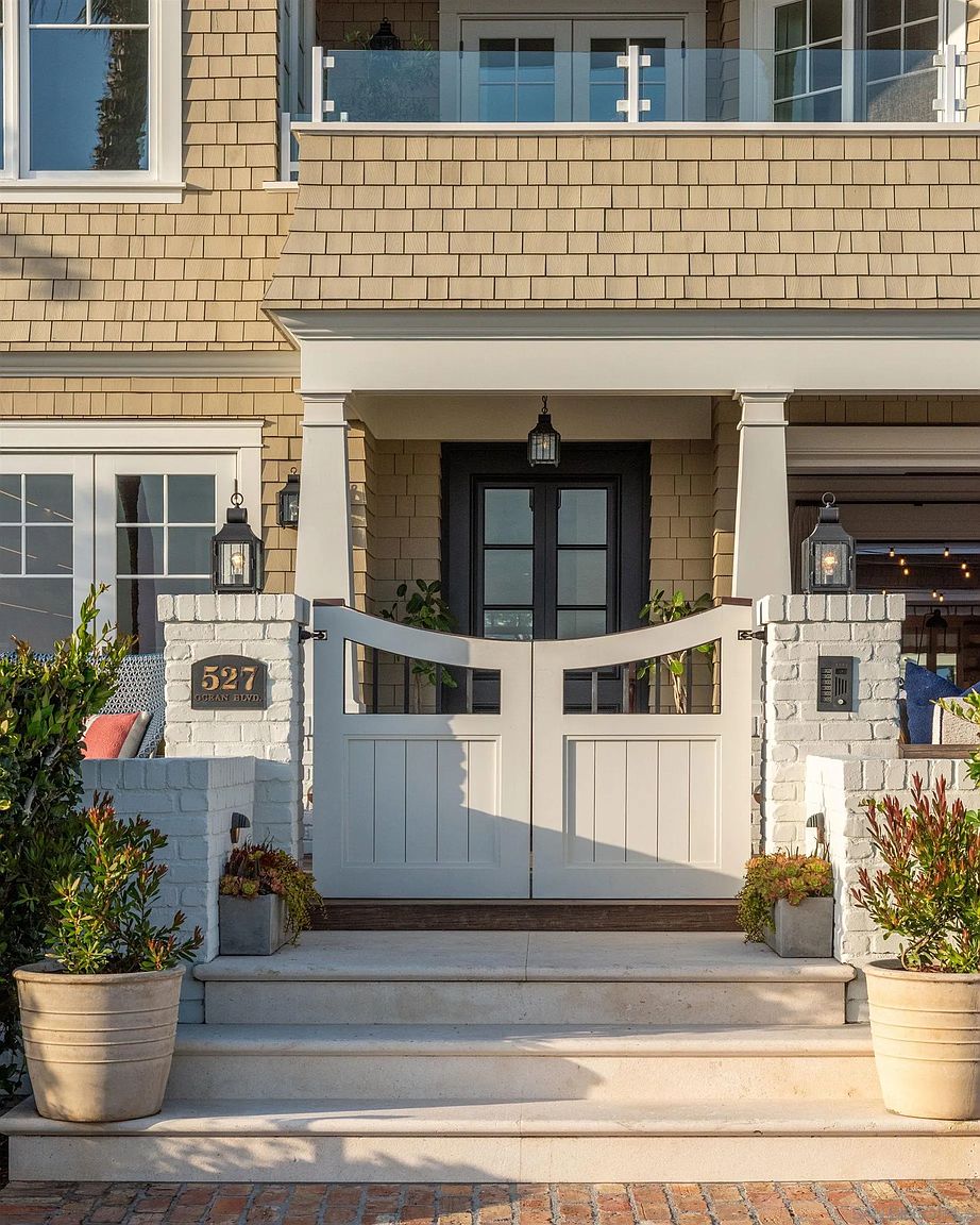 This inviting front entryway features a symmetrical design with a white double gate leading to a dark-framed front door. The facade is clad in light-toned shingles, complemented by white brick pillars topped with classic lanterns and a house number plaque. Stone steps lead up to the gate, framed by potted plants, creating a polished and welcoming curb appeal.