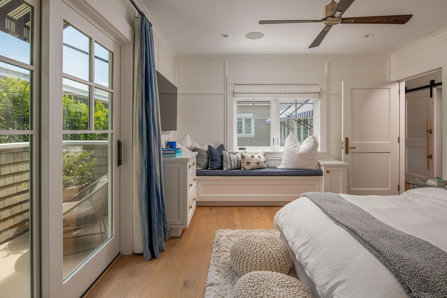 This bright and airy primary bedroom features light wood flooring, a built-in window seat with plush cushions, and a white dresser topped with a television. A glass door leads to a private balcony, while a textured rug and two knitted ottomans add warmth to the space. The room is finished with a ceiling fan and crisp white walls, creating a serene and inviting atmosphere.