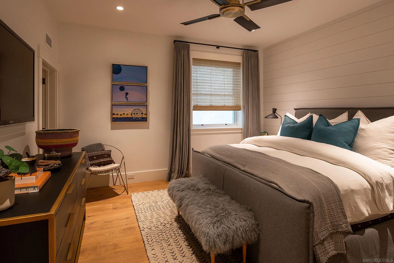 This primary bedroom features a sophisticated, modern aesthetic with a shiplap accent wall behind a plush, upholstered bed frame. The room is furnished with a textured gray bench at the foot of the bed, a sleek dresser, and a minimalist accent chair, all set against warm light wood flooring. A window with soft drapery and a woven shade allows natural light to fill the space, while a ceiling fan and wall-mounted art add functional and decorative touches.