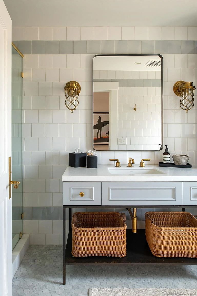 This bright and airy bathroom features a vanity with a light-toned cabinet, a stone countertop, and an integrated sink. The wall is adorned with white square tiles accented by a horizontal band of blue-grey tiles, complemented by brass-toned wall sconces and a rectangular mirror. Below the vanity, two woven baskets provide organized storage, while the floor is finished with a light-colored hexagonal tile pattern.