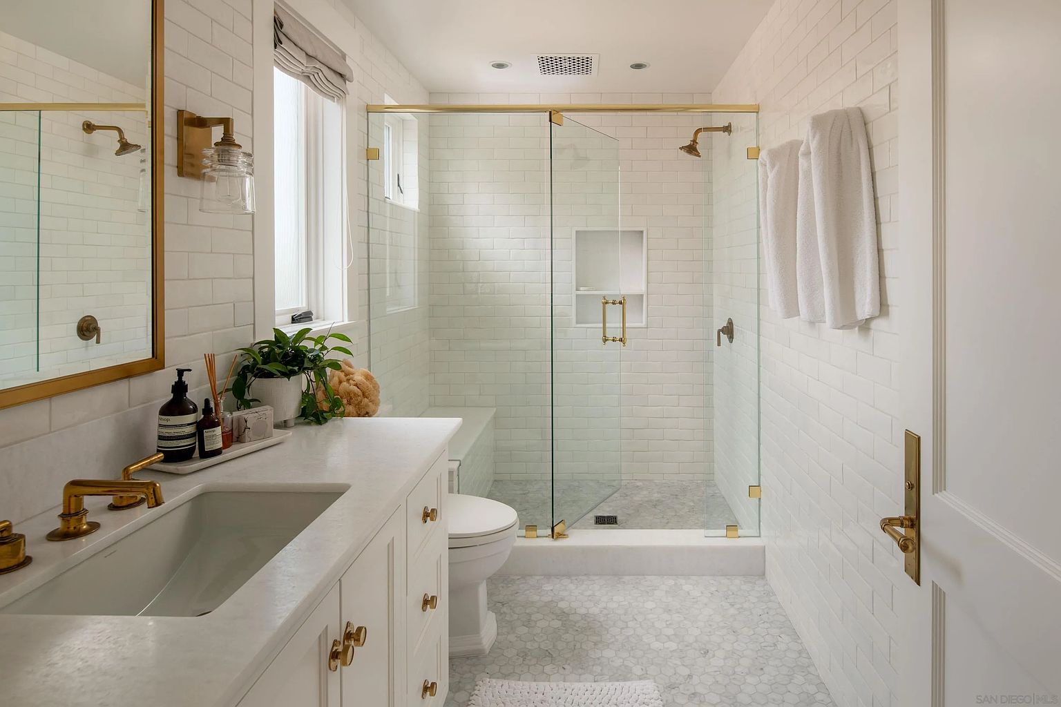 This bright and elegant bathroom features a white vanity with a stone countertop and gold-toned hardware, complemented by classic white subway tile walls. A glass-enclosed shower with a built-in niche and matching gold fixtures sits at the back of the room, adjacent to a white toilet. The space is finished with light-toned hexagonal floor tiles, creating a clean, cohesive, and sophisticated aesthetic.