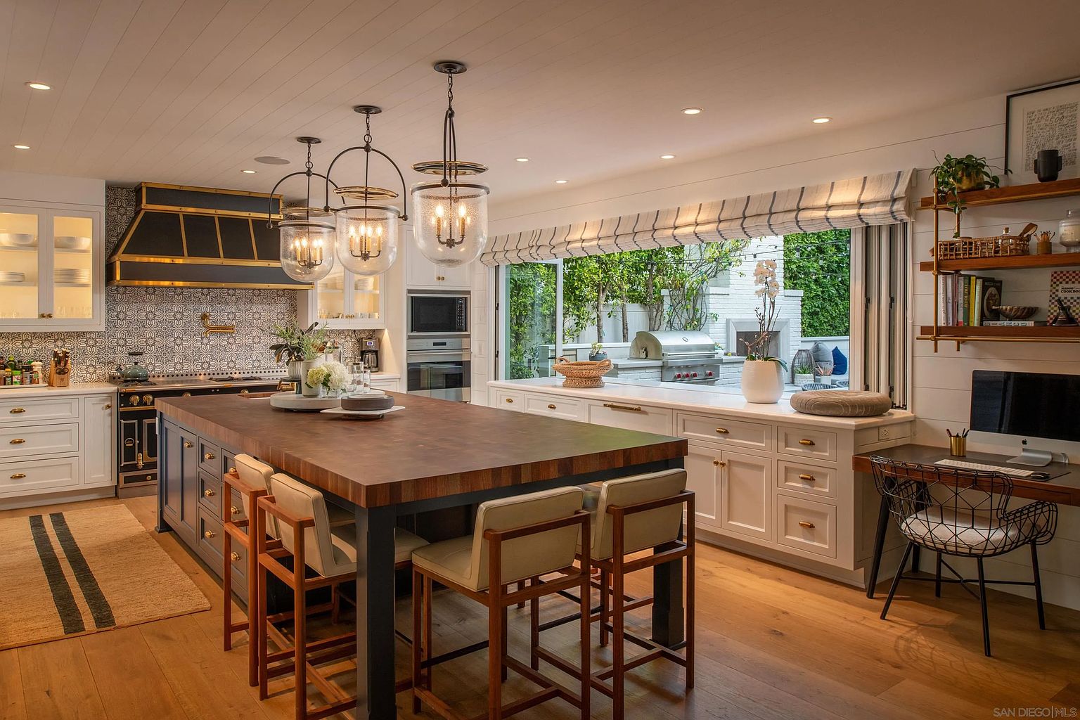 This bright and airy kitchen features a large central island with a dark wood-toned top and integrated seating, complemented by white cabinetry and a patterned tile backsplash. A prominent black and gold range hood serves as a focal point, while a wide window opening connects the indoor space to an outdoor patio area. The room is finished with light wood flooring and elegant pendant lighting, creating a sophisticated and inviting atmosphere.