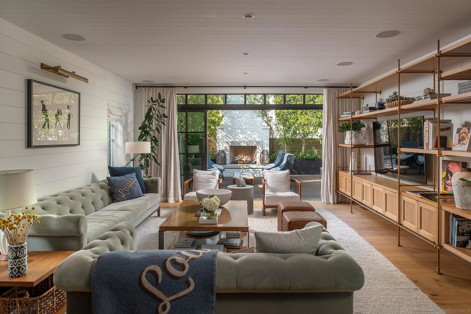 This sophisticated living room features a plush, tufted sectional sofa and a central coffee table, creating a comfortable and inviting seating area. The space is anchored by a large shelving unit with integrated lighting and a television, while a wall of glass doors opens to a private outdoor patio with a brick fireplace. The room is finished with light wood flooring, white shiplap walls, and a neutral color palette that emphasizes a bright, airy, and modern aesthetic.