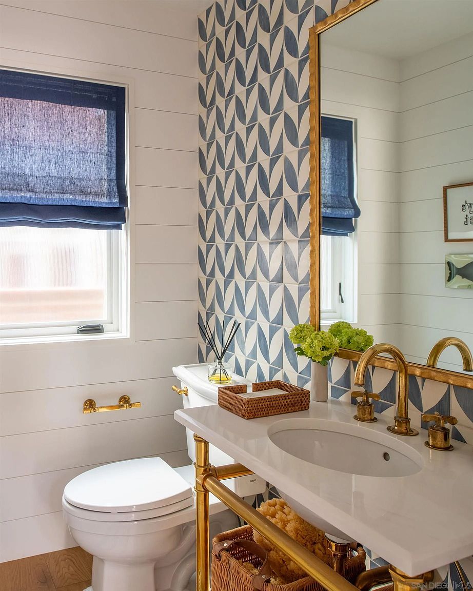 This bright and stylish powder room features a distinctive blue and white patterned tile accent wall that serves as a focal point. The space includes a white pedestal-style vanity with a gold-toned metal frame, a matching gold faucet, and a large mirror reflecting the room's shiplap-style wall paneling. A window with a dark blue roman shade provides natural light, while a woven basket under the vanity adds a touch of texture to the clean, coastal-inspired design.