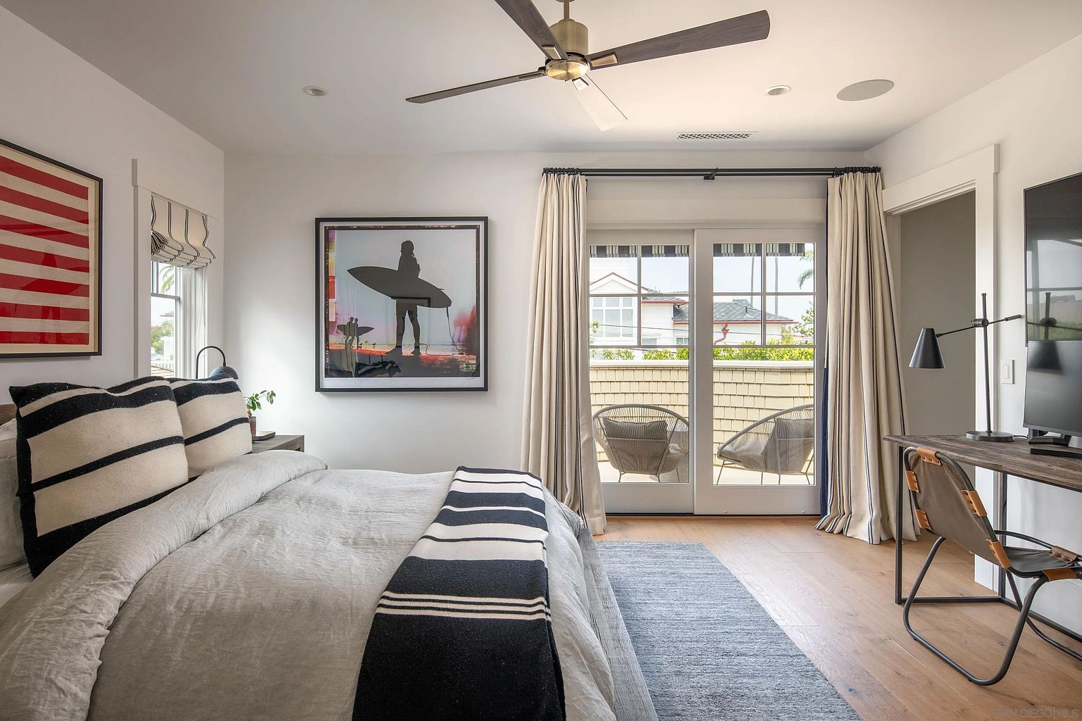 This bright and airy primary bedroom features a neutral color palette with coastal-inspired decor, including a bed dressed in striped linens and a large surf-themed art piece. A desk area is positioned to the right, while glass doors with floor-to-ceiling curtains open onto a private balcony furnished with two chairs. The room is completed by light wood flooring, a modern ceiling fan, and a large window with striped roman shades.